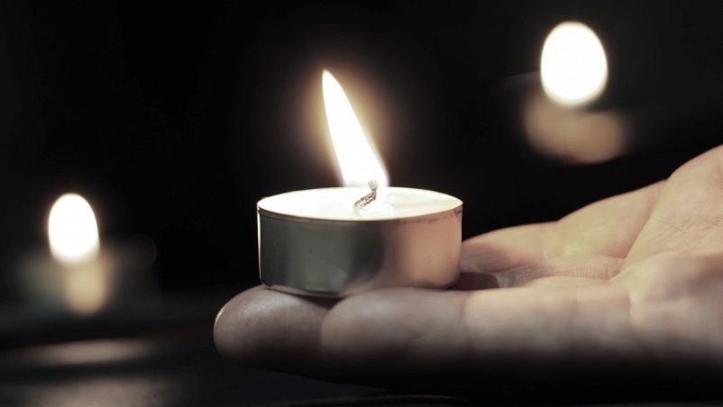 In primo piano la fiamma di un lume posato sul palmo di una mano, sullo sfondo luci di candele sfocate: Giornata della Memoria, commemorazione della Shoah